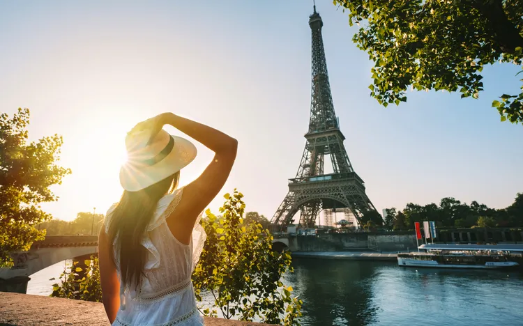 Touristin mit Sonnenhut und weißem Kleid steht bei Sonnenuntergang vor dem Eiffelturm in Paris