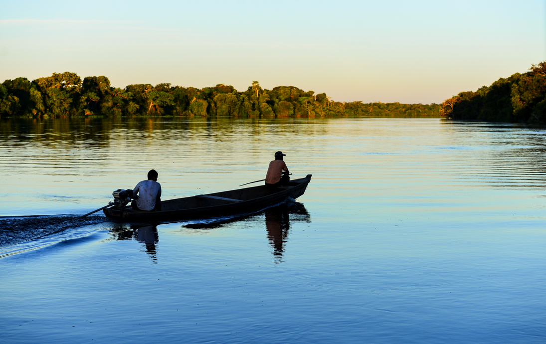 zwei Männer auf einem Boot bei Sonnenuntergang auf dem von Regenwald gesäumten Fluss Guaporé, in der Nähe des abgelegenen Dorfes Mateguá an der Grenze zu Rondônia, Brasilien - © Pedro - stock.adobe.com